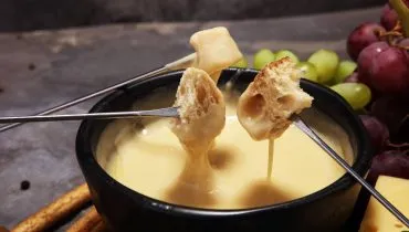 Imagem mostra um perfeito Fundue de queijo caseiro, com pães e frutas ao fundo.