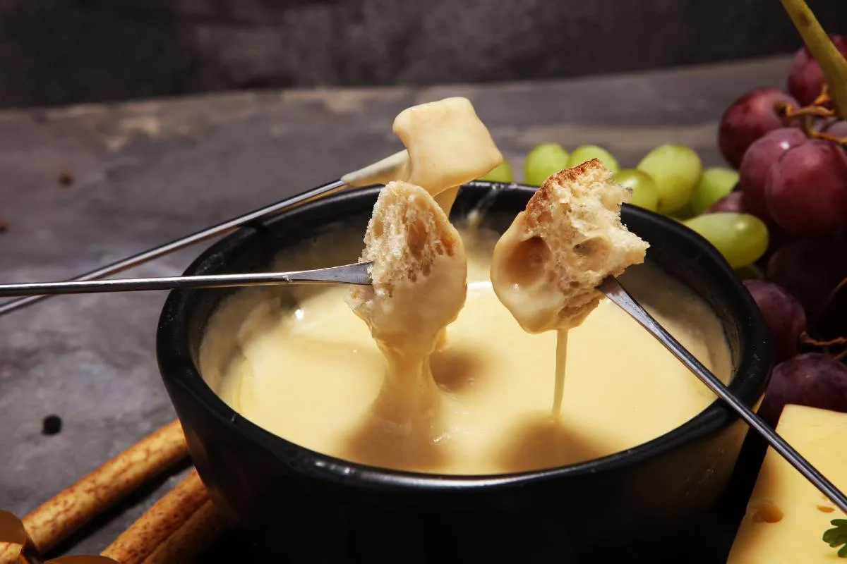 Imagem mostra um perfeito Fundue de queijo caseiro, com pães e frutas ao fundo.