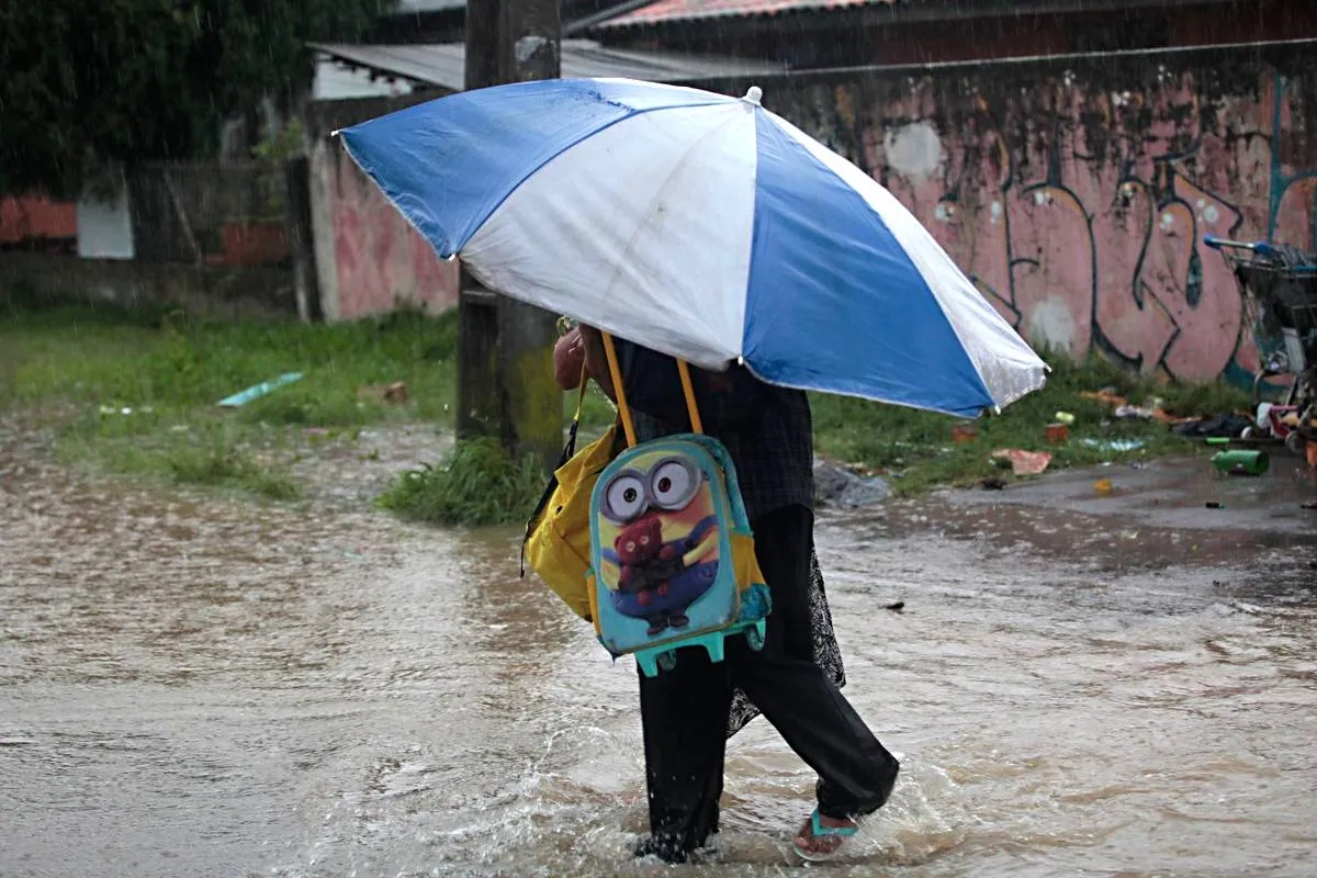 Imagem mostra uma pessoa em uma enchente com um guarda sol com as cores azul e branco e uma mochila escolar de criança.