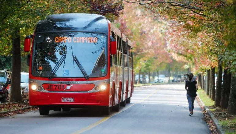 Imagem mostra um ônibus biarticulado vermelho em uma rua de Curitiba com árvores com folhas secas.