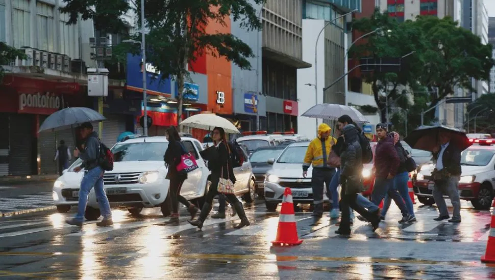 Imagem mostra um dia de chuva e céu fechado em Curitiba, com pessoas de guarda-chuva passando em uma rua bem escura por causa do tempo fechado.