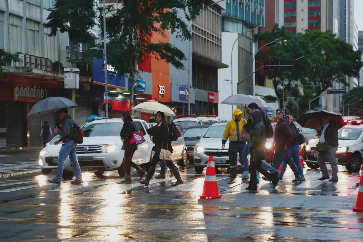 Imagem mostra um dia de chuva e céu fechado em Curitiba, com pessoas de guarda-chuva passando em uma rua bem escura por causa do tempo fechado.