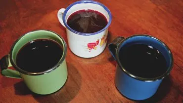 Imagem mostra três canecas com quentão dispostas em uma mesa de madeira. Uma delas é verde, outra azul e a terceira é branca.