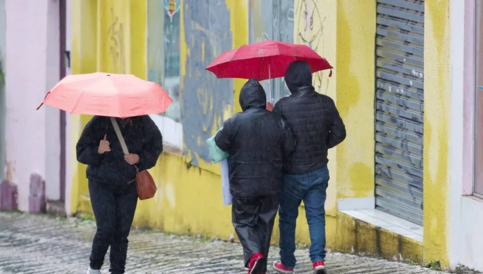 Imagem mostra três pessoas andando em um dia de chuva, com guarda chuva vermelho e rosa.