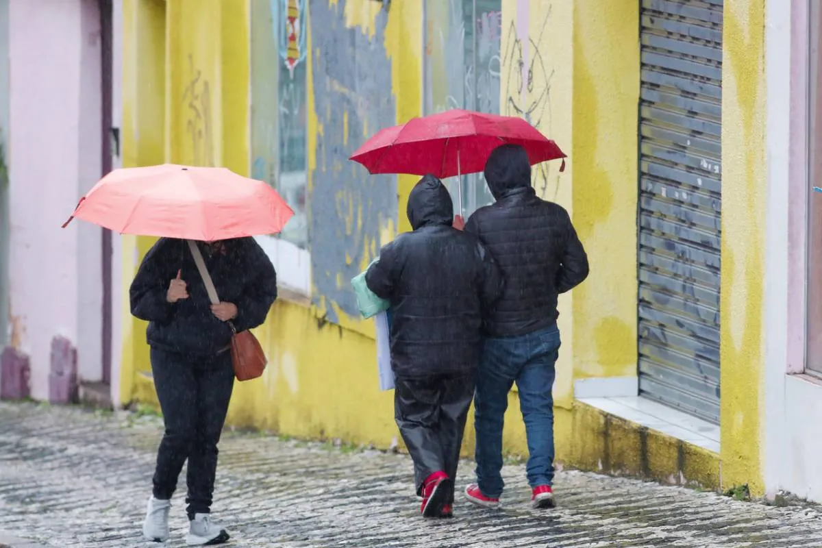 Imagem mostra três pessoas andando em um dia de chuva, com guarda chuva vermelho e rosa.