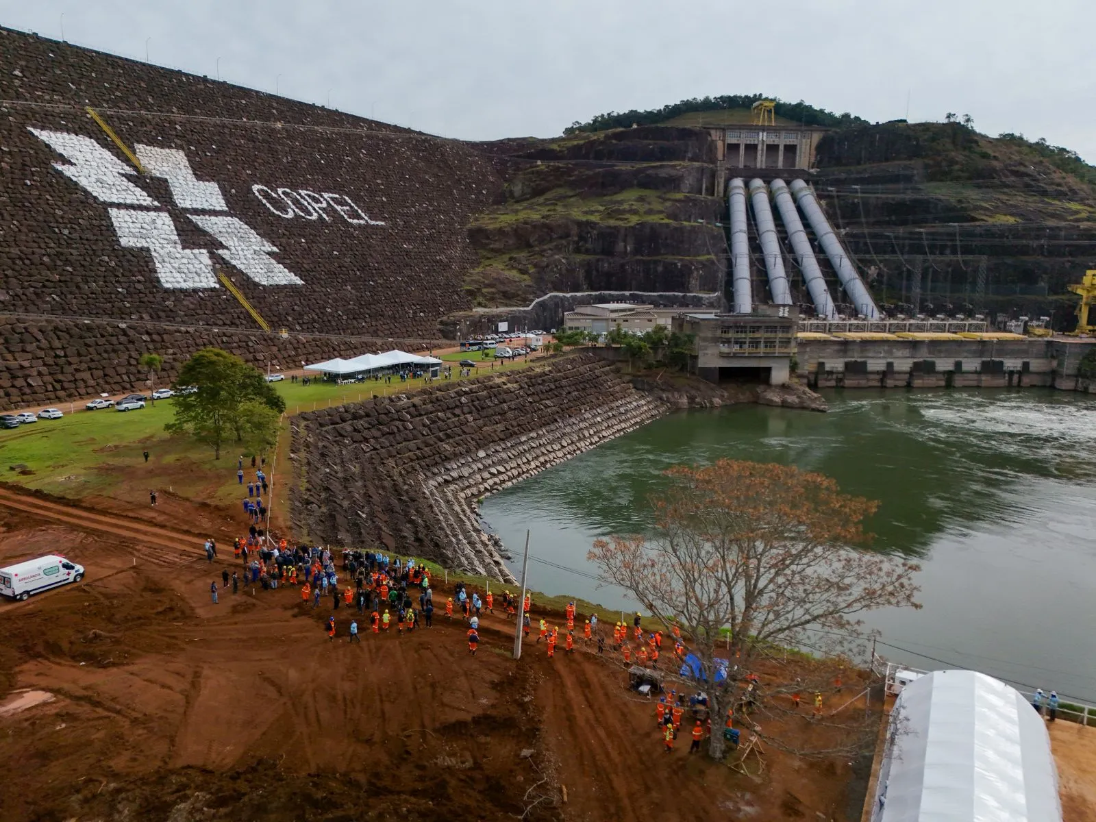 Imagem feita de longa distância mostra a estrutura de concreto da barragem da Usian de Segredo, com o leito do rio à direita. No centro da imagem, bem distante, centenas de operários e autoridades que participaram do lançamento