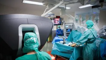 Na imagem um médico utiliza um equipamento moderno com tela e comandos para operar um robo que executa uma cirurgia ao fundo