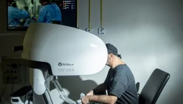 Na imagem um médico utiliza um equipamento moderno com tela e comandos para operar um robo que executa uma cirurgia ao fundo