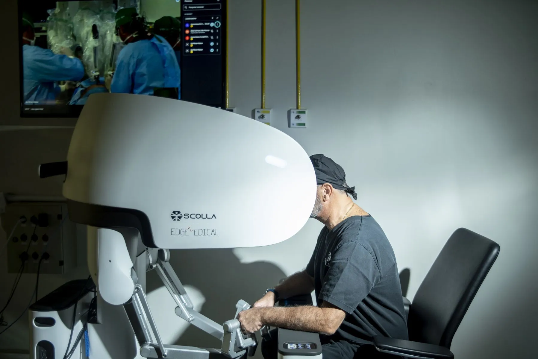 Na imagem um médico utiliza um equipamento moderno com tela e comandos para operar um robo que executa uma cirurgia ao fundo