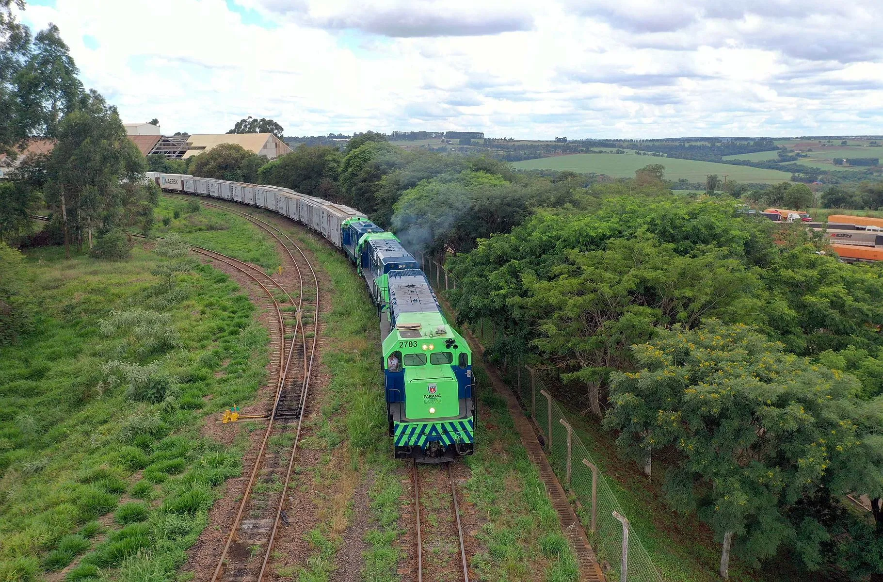 Imagem aérea mostra locomotiva puxando vários vagões, numa curva, com caminhões estacionados à direita e muitas árvores formando o corredor