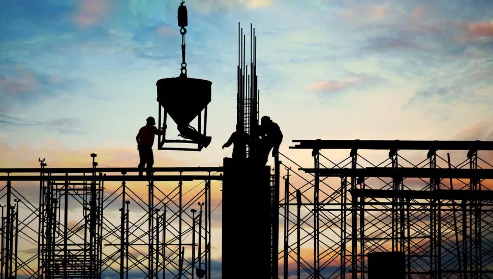 Foto mostra um contraluz com final de tarde ao fundo, e em primeiro plano andaimes de obras, uma grande coluna ao centro, com três operários conduzindo um dispensador de concreto.