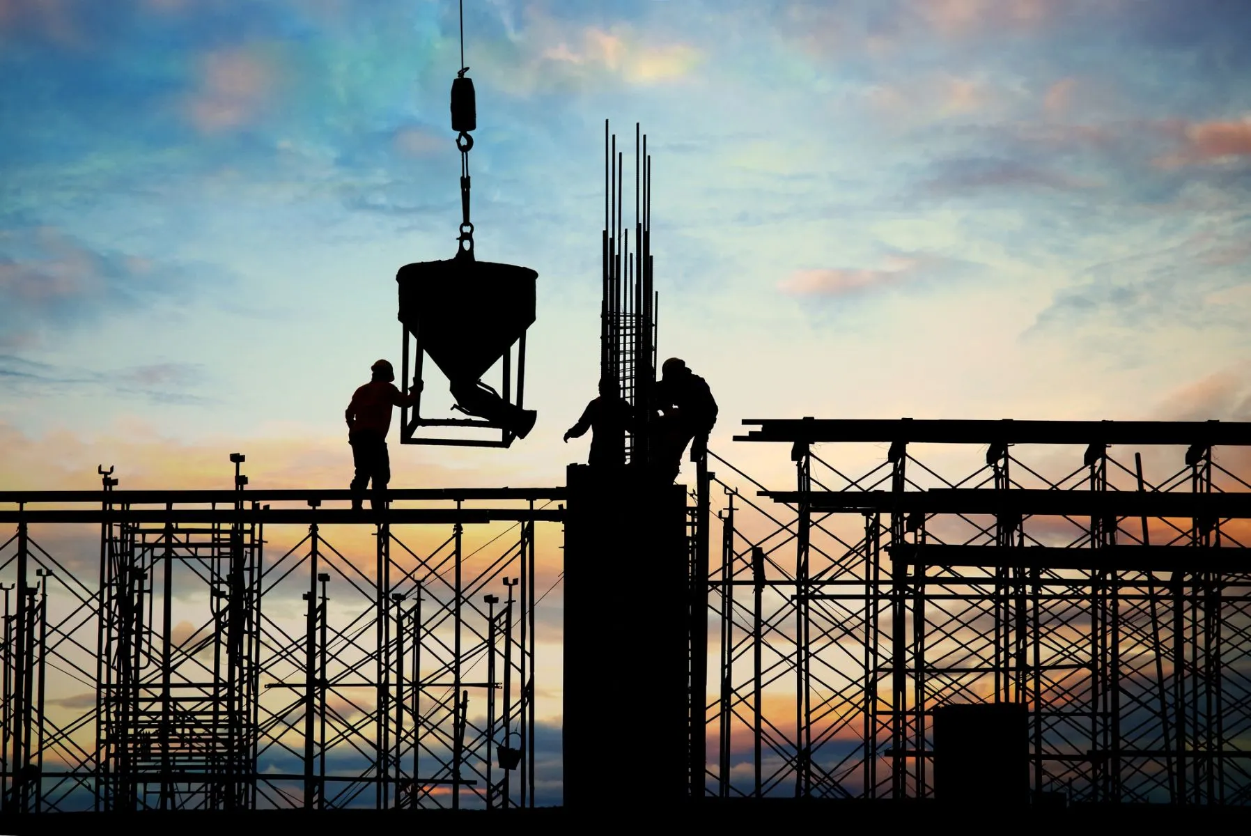 Foto mostra um contraluz com final de tarde ao fundo, e em primeiro plano andaimes de obras, uma grande coluna ao centro, com três operários conduzindo um dispensador de concreto.