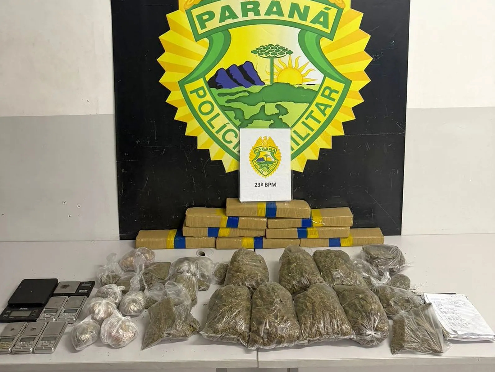 Imagem mostra vários pacote de droga espalhados numa mesa, as balanças apreendidas, com um banner da Polícia Militar ao fundo