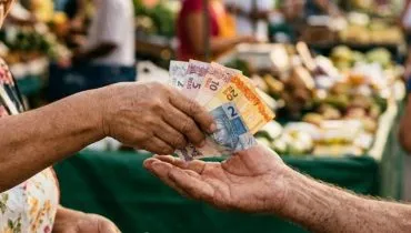 Uma imagem de close-up foca nas mãos centralizadas de uma feirante de meia-idade e de um cliente idoso em uma feira de rua brasileira ao ar livre e movimentada. A feirante, com um avental floral, está colocando várias notas de Real brasileiro usadas de várias denominações (visíveis, mas ligeiramente desfocadas) diretamente na palma aberta e envelhecida do cliente. Suas mãos estão entrelaçadas, centralizadas na composição. A feirante olha para a transação com um sorriso gentil, enquanto o cliente idoso olha com uma expressão de satisfação. O fundo é um borrão quente e vibrante de atividades do mercado, barracas desfocadas com frutas e vegetais, outras pessoas e a luz do sol da tarde. A fotografia documental e autêntica tem grãos visíveis e profundidade de campo muito rasa, focando apenas no dinheiro e no ponto de contato das mãos. A iluminação é natural e quente.