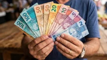 Esta foto foca na textura e na diversidade de cores das notas. Uma mão (bem cuidada, com unhas curtas) segura um leque perfeito e denso de notas. Estão visíveis todas as denominações principais (R$2, R$5, R$10, R$20, R$50, R$100 e R$200). O fundo é um mercado de rua brasileiro desfocado (bokeh), trazendo um contexto real de uso do dinheiro. A luz é natural e suave.