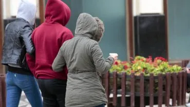 Três pessoas são vistas de costas caminhando em um ambiente urbano. À esquerda, uma pessoa usa capuz cinza e jaqueta de couro preta; no centro, uma pessoa veste um moletom vermelho com capuz; à direita, a terceira pessoa usa uma jaqueta verde-oliva com capuz de detalhe em pele sintética e segura um copo de bebida quente na mão esquerda. Ao fundo, há uma floreira de madeira com flores vermelhas e a fachada texturizada de um edifício. A atmosfera sugere um clima frio e nublado.