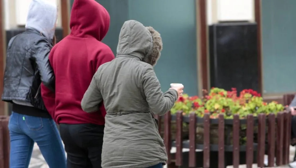 Três pessoas são vistas de costas caminhando em um ambiente urbano. À esquerda, uma pessoa usa capuz cinza e jaqueta de couro preta; no centro, uma pessoa veste um moletom vermelho com capuz; à direita, a terceira pessoa usa uma jaqueta verde-oliva com capuz de detalhe em pele sintética e segura um copo de bebida quente na mão esquerda. Ao fundo, há uma floreira de madeira com flores vermelhas e a fachada texturizada de um edifício. A atmosfera sugere um clima frio e nublado.