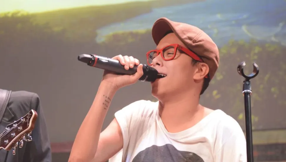 Imagem mostra a cantora Maria Gadu. Ela está cantando no microfone, com uma camiseta branca e uma boina bege.
