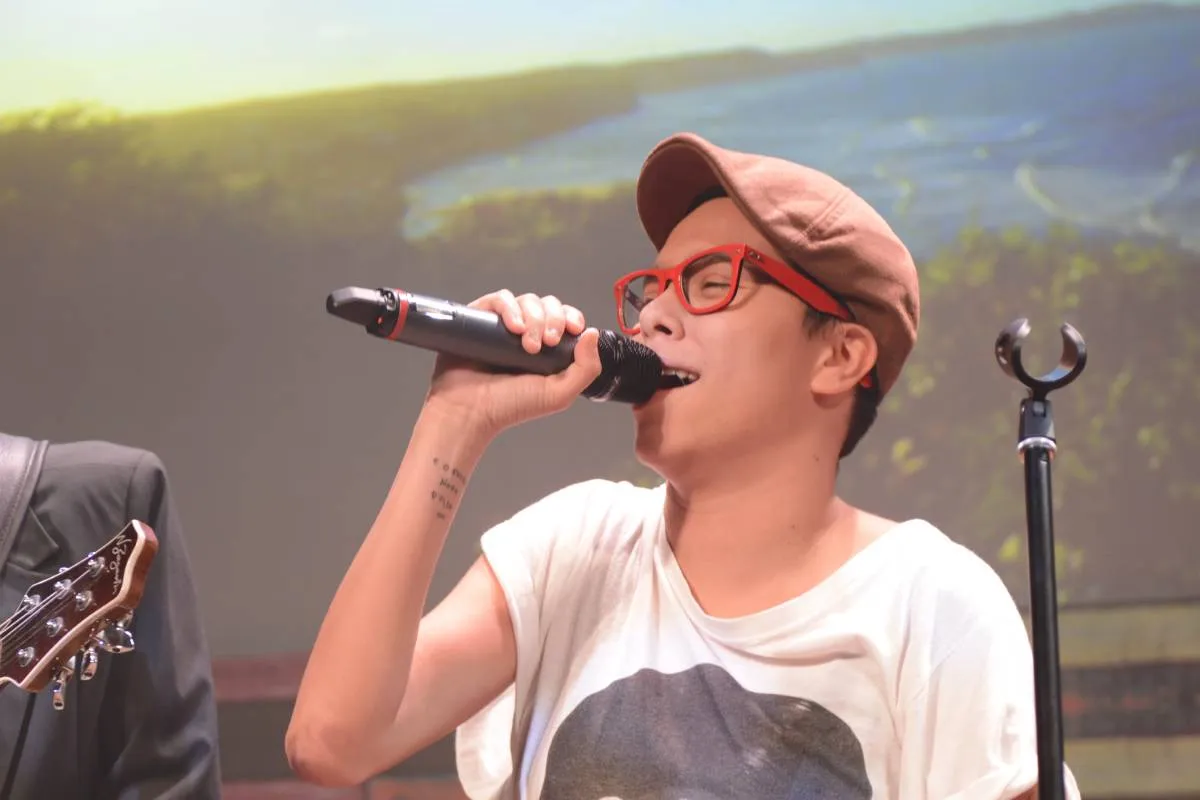 Imagem mostra a cantora Maria Gadu. Ela está cantando no microfone, com uma camiseta branca e uma boina bege.