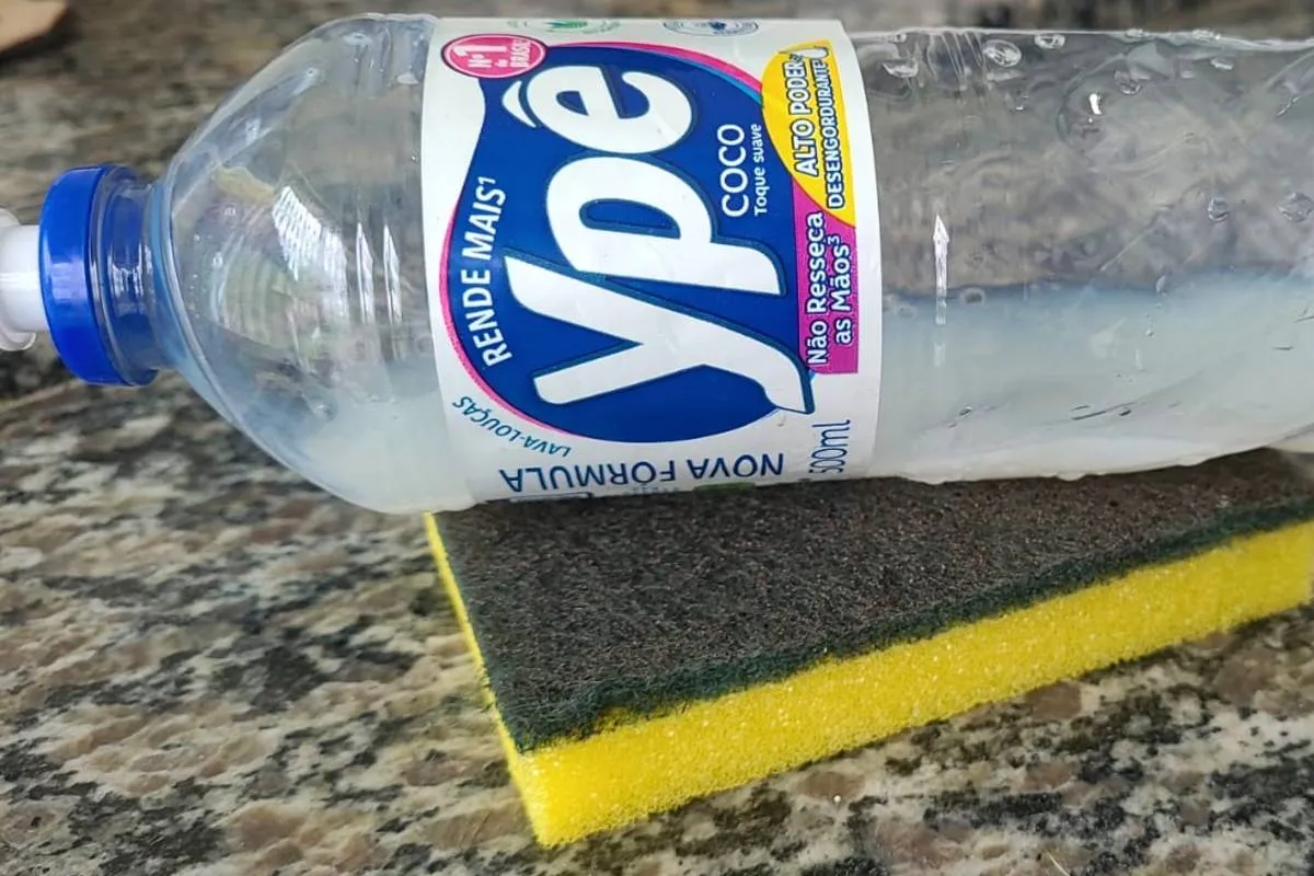 Imagem mostra um frasco plástico de detergente líquido Ypê Coco deitado sobre uma esponja de lavar louça amarela e verde.
