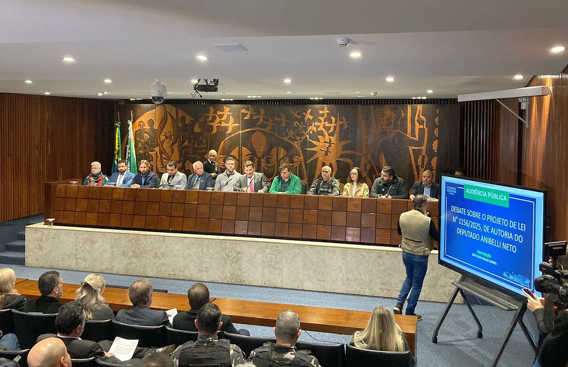 imagem mostra convidados sentados à uma mesa, num palco, com vários convidados na plateia, num auditório. Um banner anuncia o tema do encontro