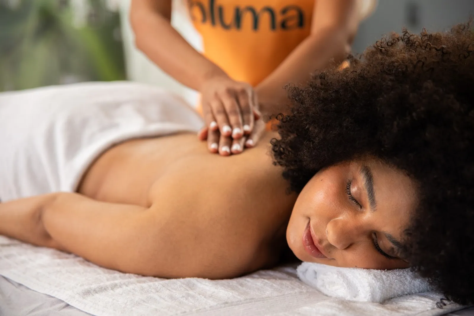 Mulher deitada de barriga para baixo em maca recebendo massagem nas costas, feita por pessoa com blusa laranja escrito Bluma em preto. Bluma é parceira da Natura.