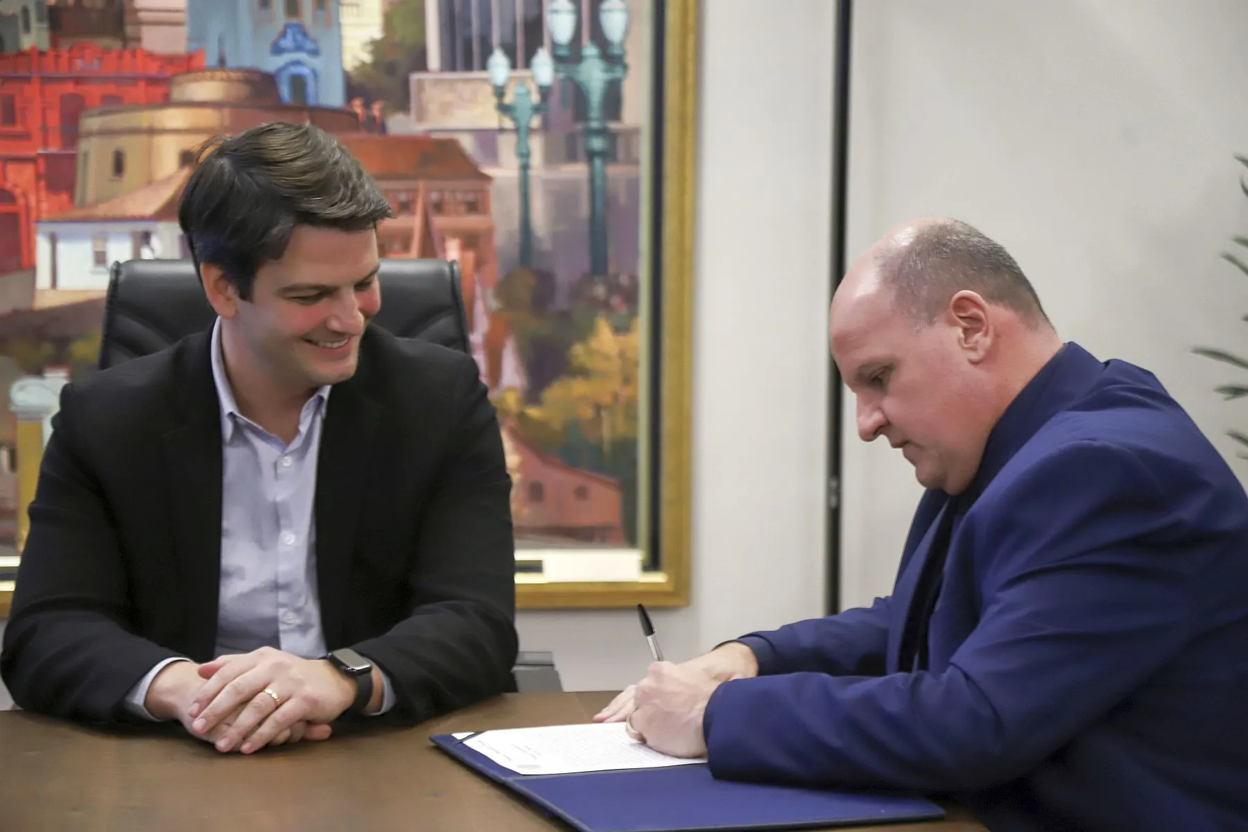 Foto mostra dois homens, um a esquerda, de terno escuro, é o prefeito Eduardo Pimentel. O outro é o vereador Leonidas Dias, de terno azul, assinando um documento