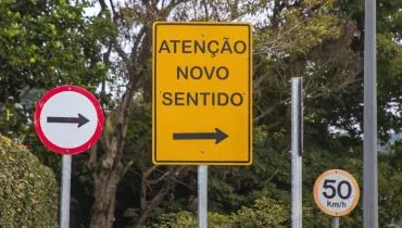 Placas de trânsito indicam novo sentido de circulação à direita em rua arborizada de Curitiba.