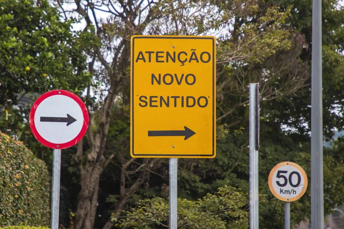 Placas de trânsito indicam novo sentido de circulação à direita em rua arborizada de Curitiba.