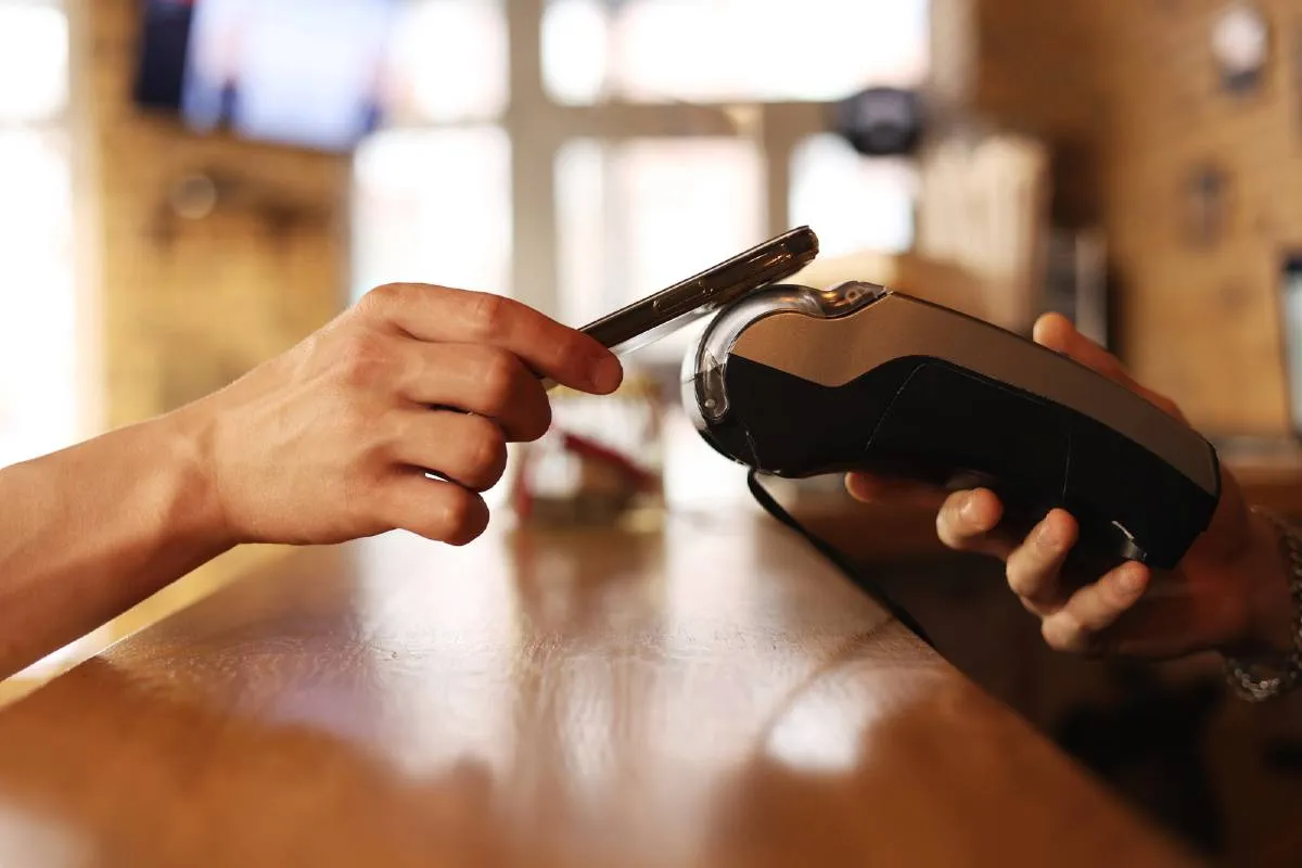 Pessoa aproximando um smartphone da tela de uma maquininha de cartão sobre um balcão de madeira.