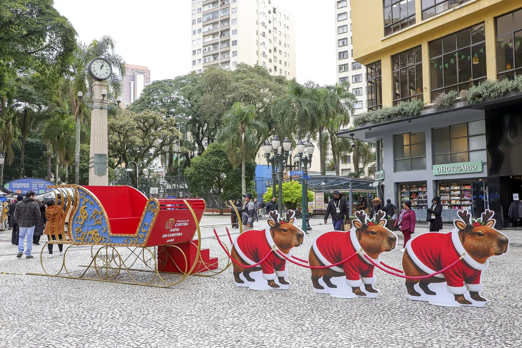 Imagem mostra um trenó vermelho, puxado por três capivaras/renas, com a praça osório ao fundo