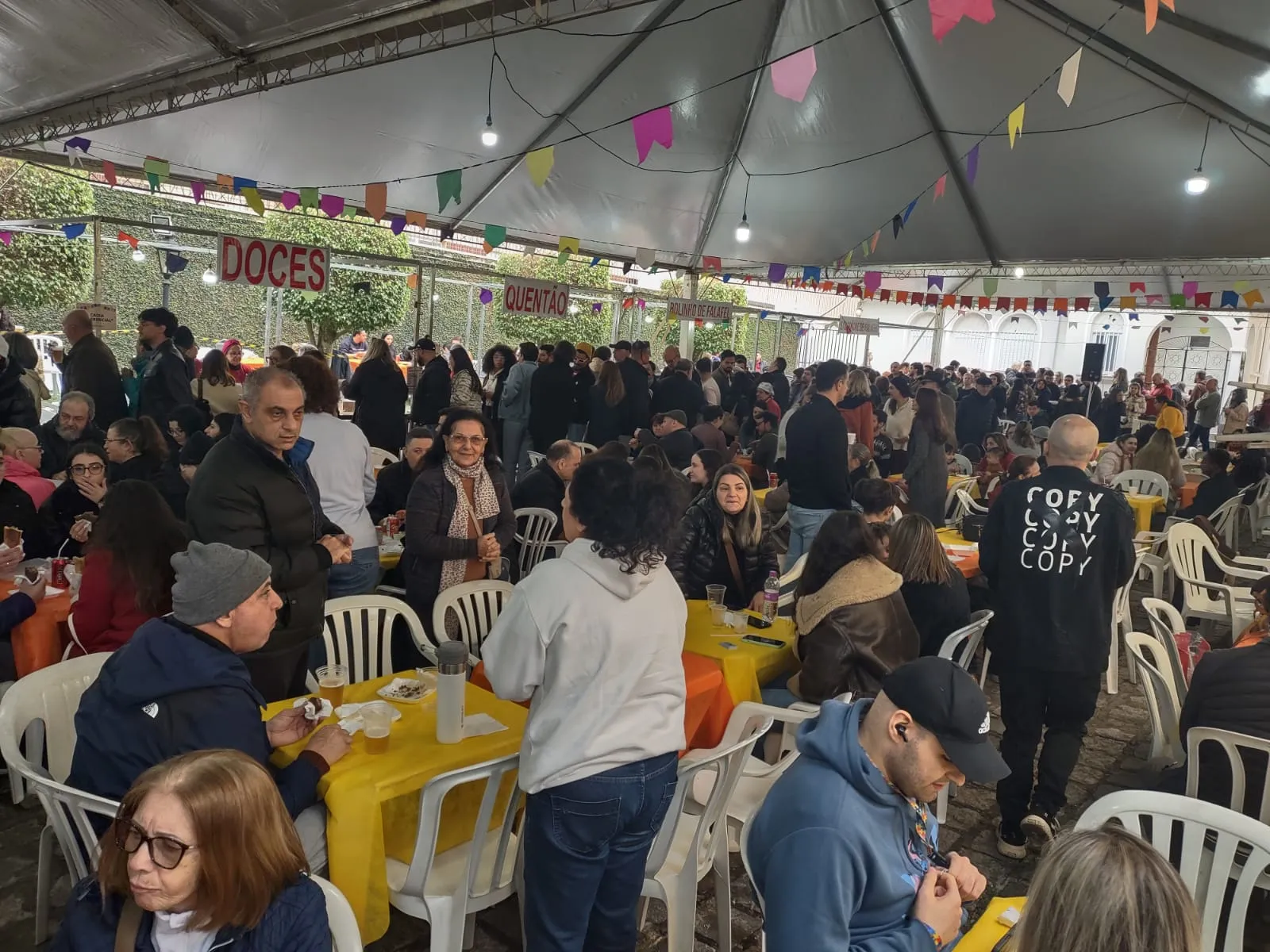Pessoas sentadas e em pé embaixo de tenda em Festa Junina da Igreja Ortodoxa