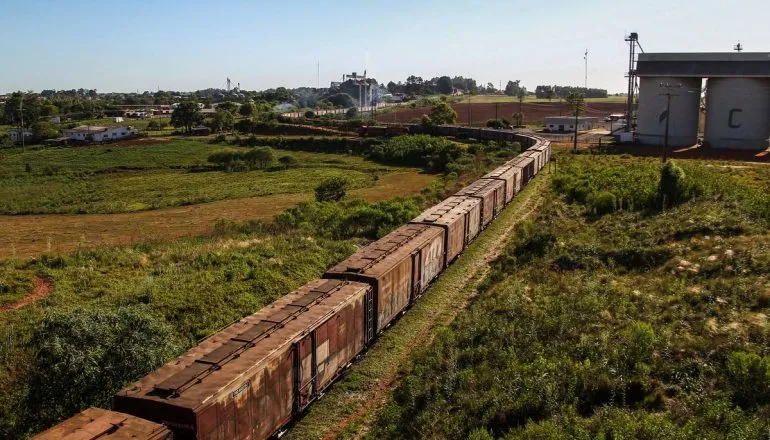 Trem de carga longo e enferrujado transportando vagões fechados por uma ferrovia que faz uma curva acentuada em uma área rural, próximo a grandes silos de armazenamento de grãos.