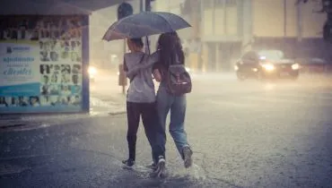 Imagem mostra uma mulher e uma criança atravessando a rua alagada, sob forte chuva, debaixo de um guarda-chuvas. Ao fundo, carros com faróis ligados