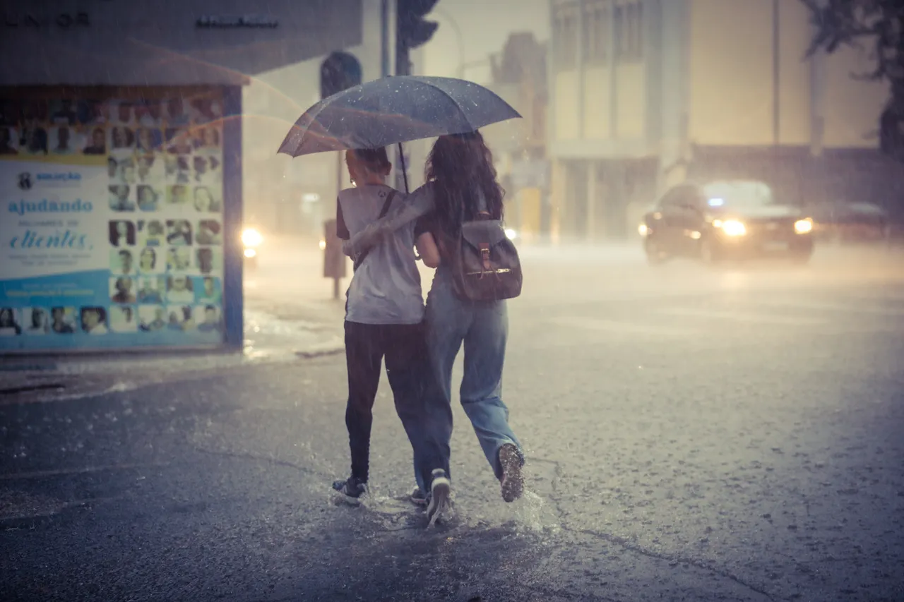 Imagem mostra uma mulher e uma criança atravessando a rua alagada, sob forte chuva, debaixo de um guarda-chuvas. Ao fundo, carros com faróis ligados