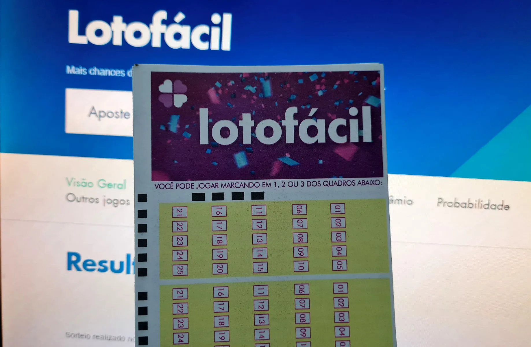 Volante de aposta da Lotofácil na frente de um monitor com resultado online da loteria