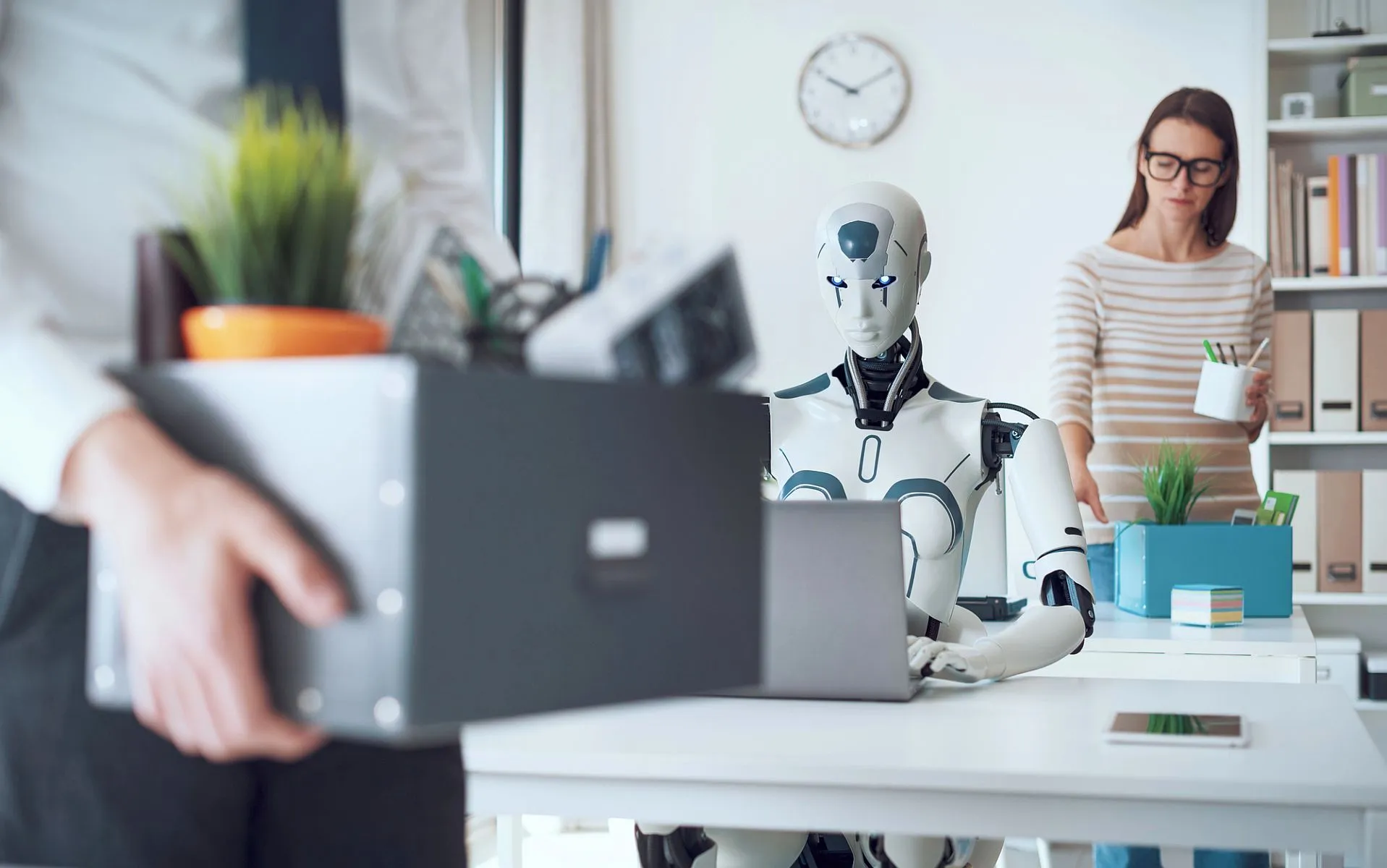 Imagem ilustrativa mostra um robo sentado numa mesa de trabalho, com um homem levando uma caixa com pertences que seriam do seu escritório antes de perder a vaga para o robo