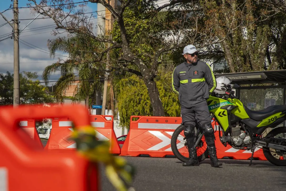 Imagem mostra um agente de trânsito ao lado de uma moto e cones de sinalização em Curitiba.