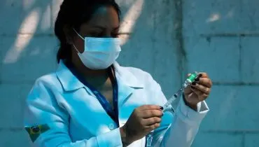 Imagem mostra uma mulher com máscara e jaleco de saúde preparando uma injeção/vacina