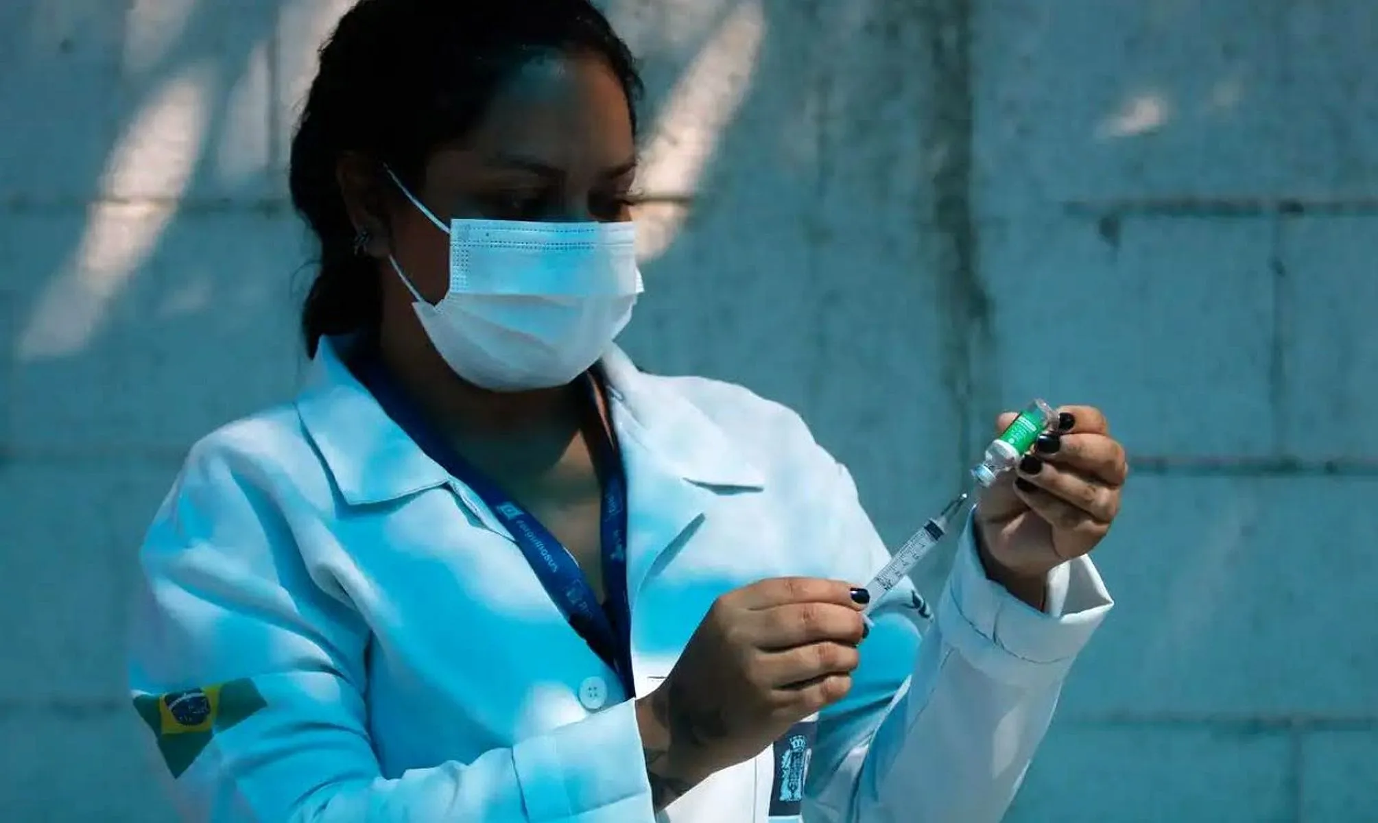 Imagem mostra uma mulher com máscara e jaleco de saúde preparando uma injeção/vacina