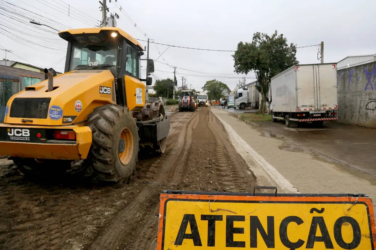 Imagem mostra uma máquina trabalhando em uma rua de Curitiba, com uma placa de "atenção" logo abaixo.