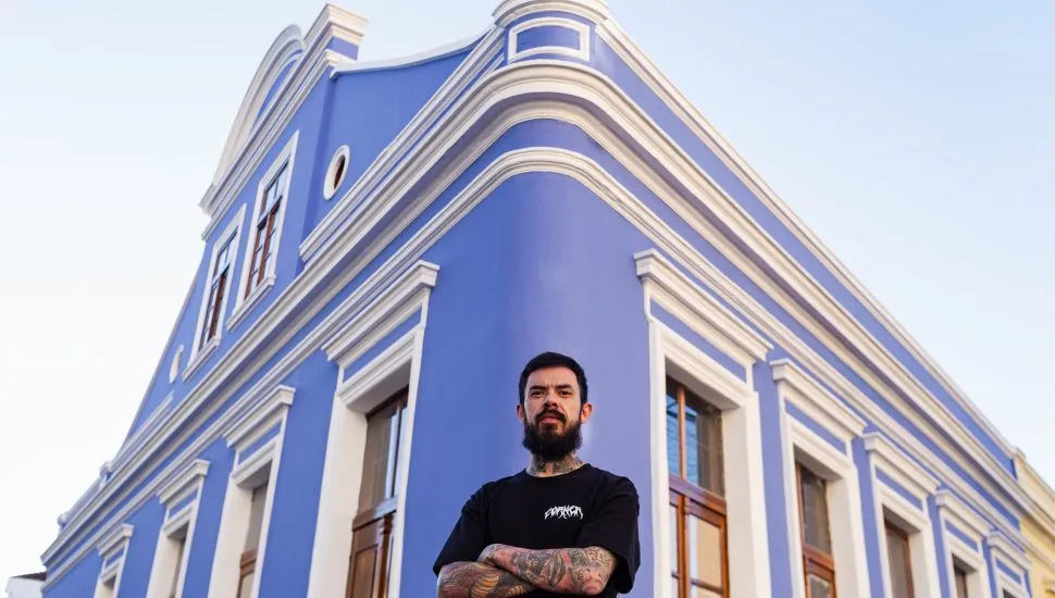Homem de barba preta, de camiseta preta, de braços cruzados em frente à Casa Maleski, casarão histórico de esquina, com paredes azuis e molduas de janelas brancas