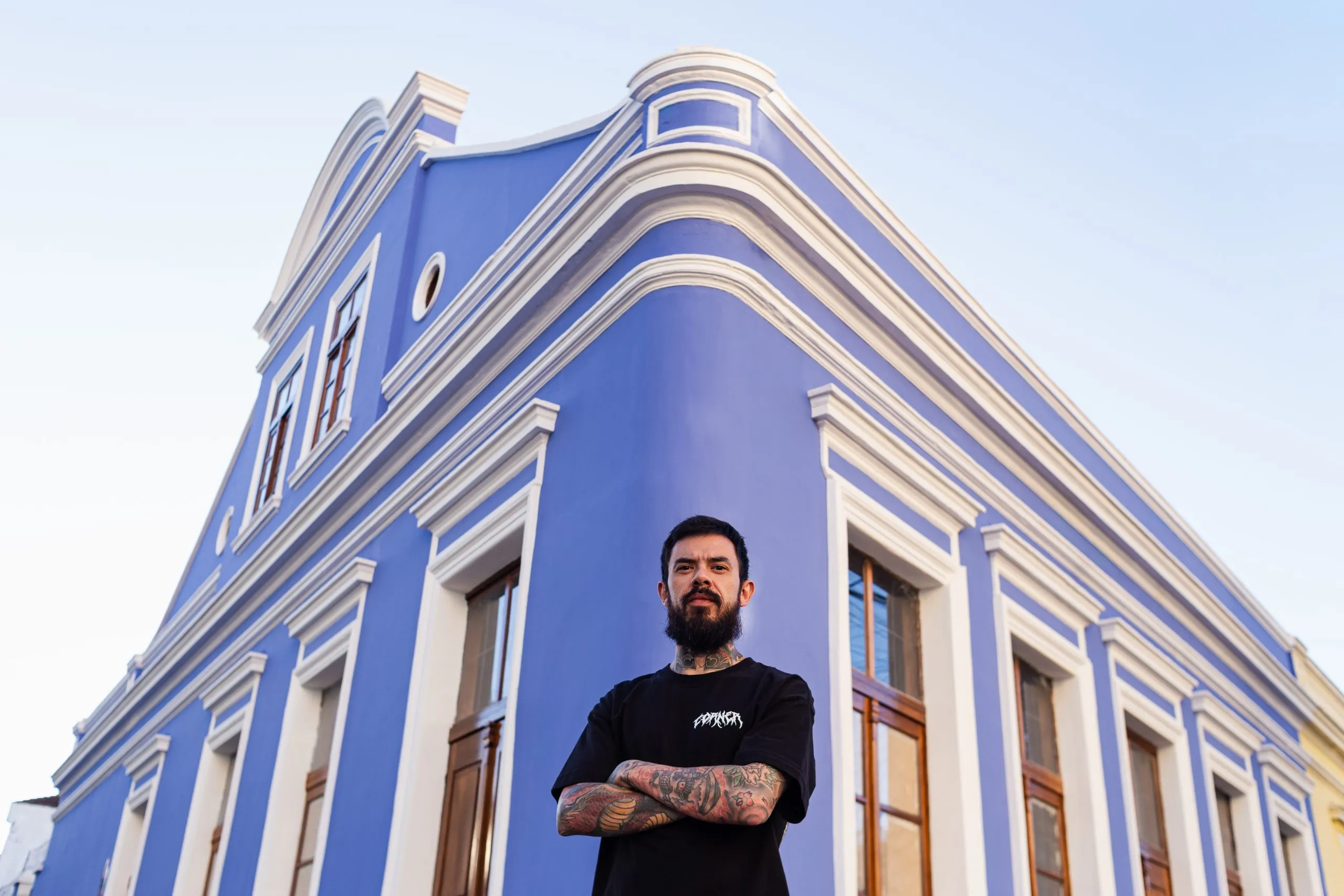 Homem de barba preta, de camiseta preta, de braços cruzados em frente à Casa Maleski, casarão histórico de esquina, com paredes azuis e molduas de janelas brancas