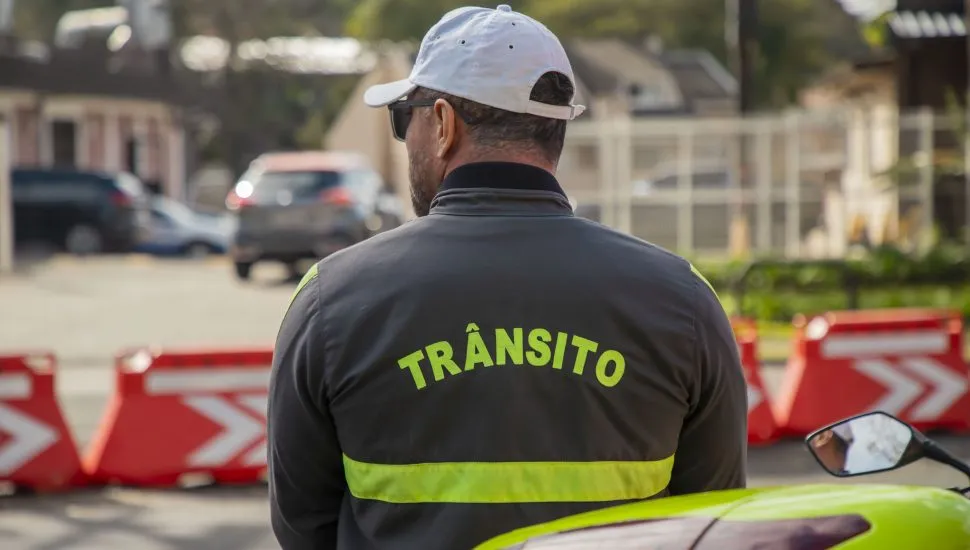 Imagem mostra um agente de trânsito na frente de cones da prefeitura de Curitiba. Ele está de costas, onde está escrito: "Trânsito"