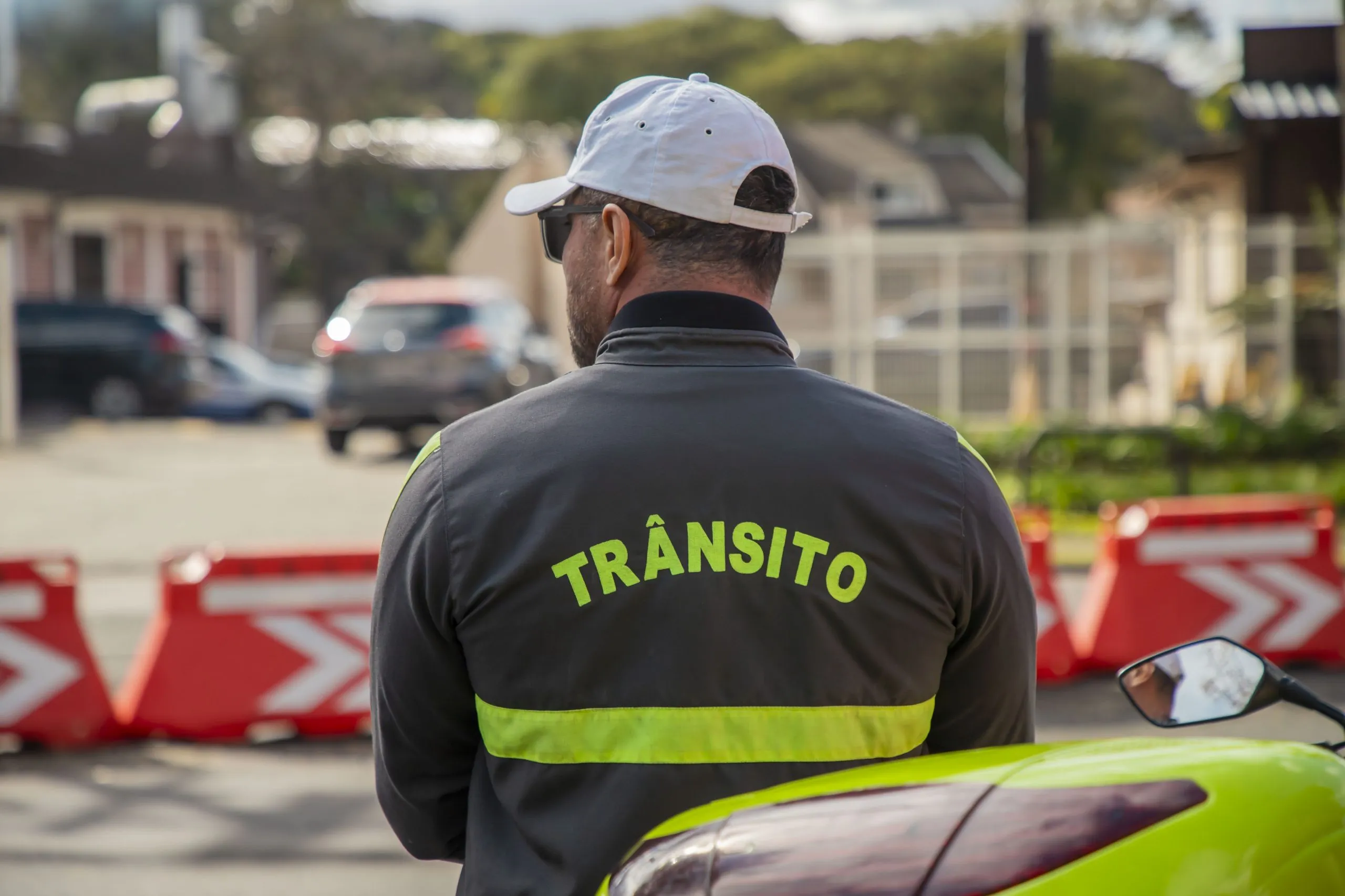Imagem mostra um agente de trânsito na frente de cones da prefeitura de Curitiba. Ele está de costas, onde está escrito: "Trânsito"