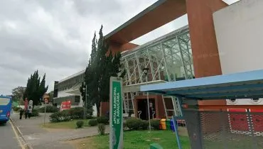 imagem mostra fachada do hospital, com placa rm primeiro plano com o nome do hospital. Ao fundo uma ambulância e na calçada pedestres