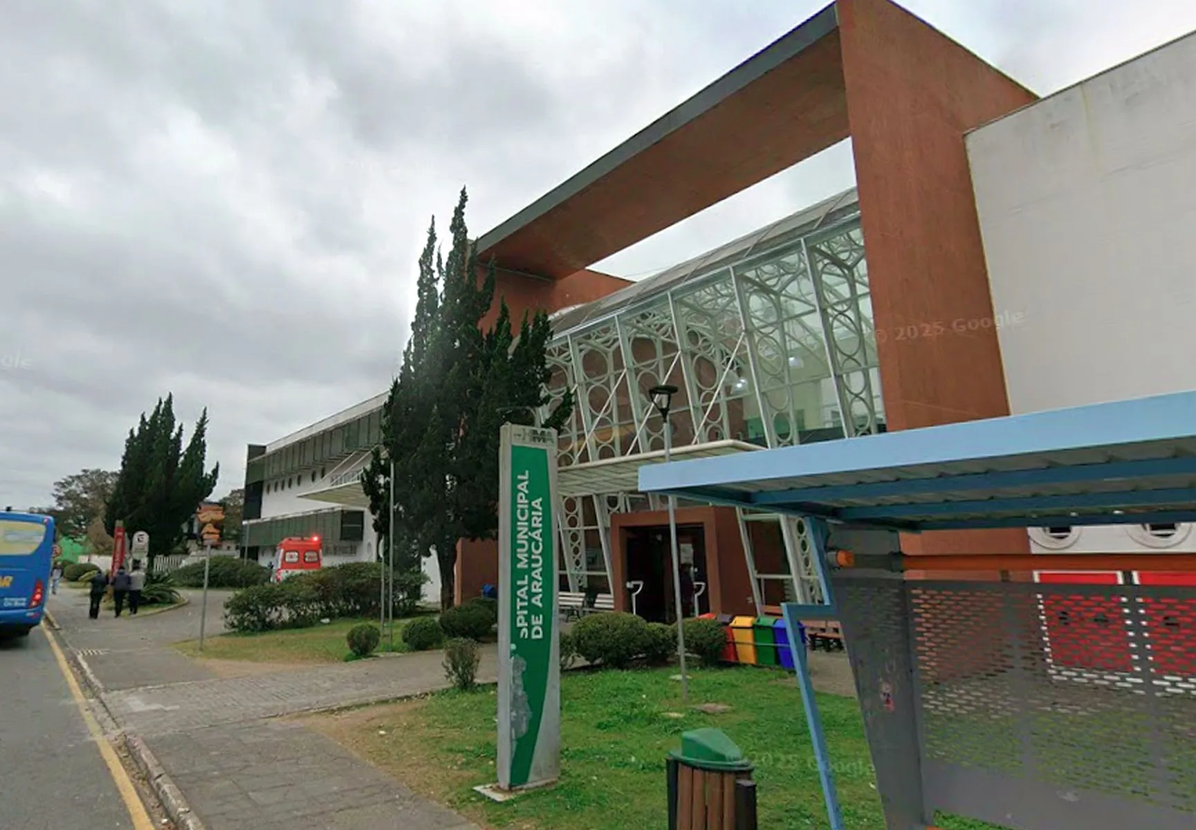 imagem mostra fachada do hospital, com placa rm primeiro plano com o nome do hospital. Ao fundo uma ambulância e na calçada pedestres