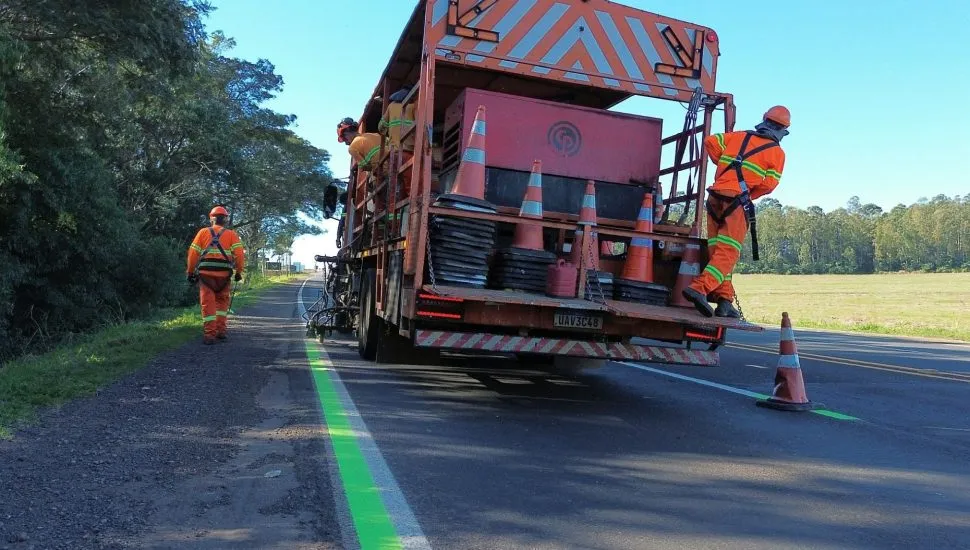 Imagem mostra caminhão com três funcionários, pintando faixas na cor verde ao lado de faixas brancas da rodovia