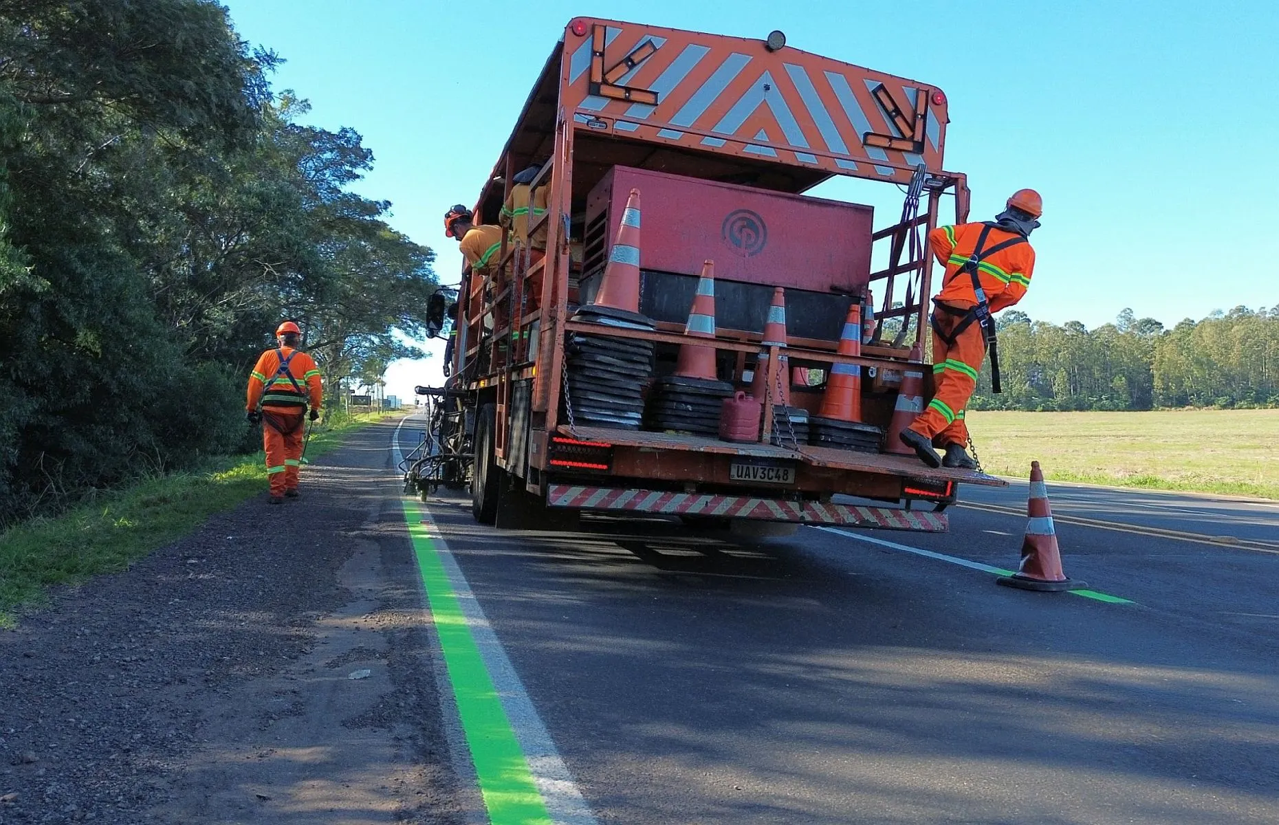 Imagem mostra caminhão com três funcionários, pintando faixas na cor verde ao lado de faixas brancas da rodovia