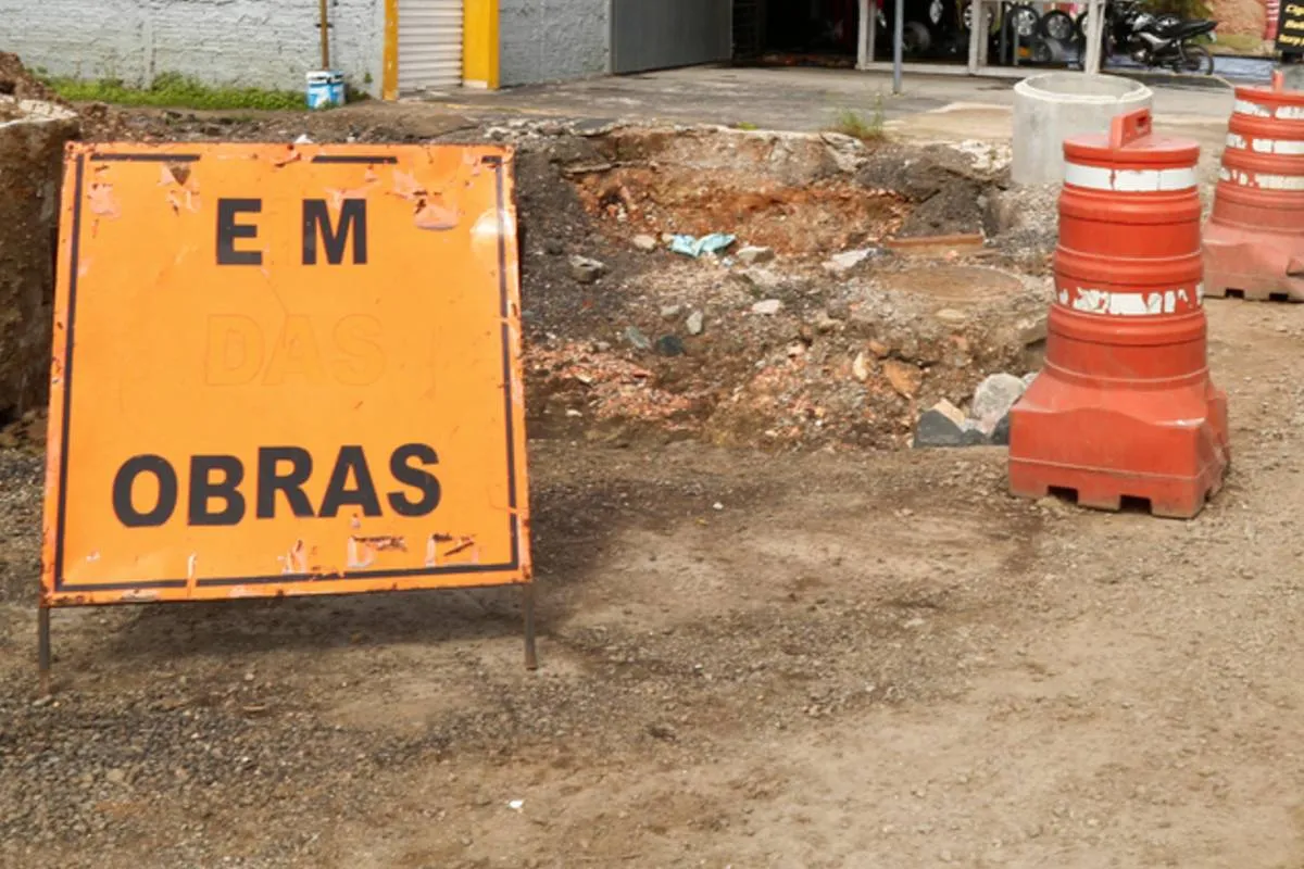 Imagem mostra uma placa de obra ao lado de um cone grande de cor laranjada.